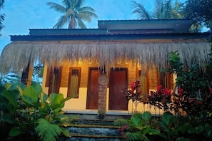 Una edificación con techo de paja, puertas y ventanas de madera, rodeada de plantas tropicales.