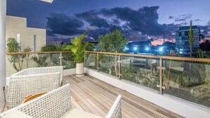 A balcony with a wooden floor, white wicker furniture, and a glass railing overlooking a cityscape at dusk. - sol N1 · Incredible 4 Bds villa near Finns club (Bali)