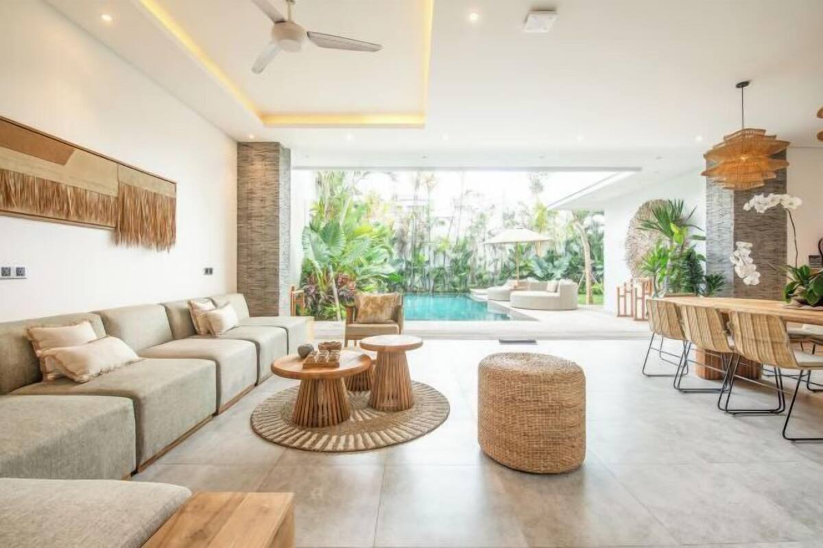 A modern living area with a sofa set, round wooden tables, a dining table with chairs, and a view of a swimming pool.