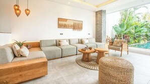 A modern living room with a sofa, coffee table, and wicker chair, a ceiling fan, and a view of a swimming pool. - sol N1 · Incredible 4 Bds villa near Finns club (Bali)