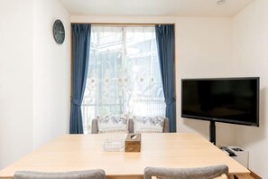 A room with a wooden dining table, two grey chairs, a flat-screen TV mounted on the wall, and a window with blue curtains. - lami home (Tokyo)