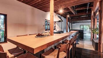 A long wooden dining table with chairs, a kitchen in the background, and a view of the outdoors.