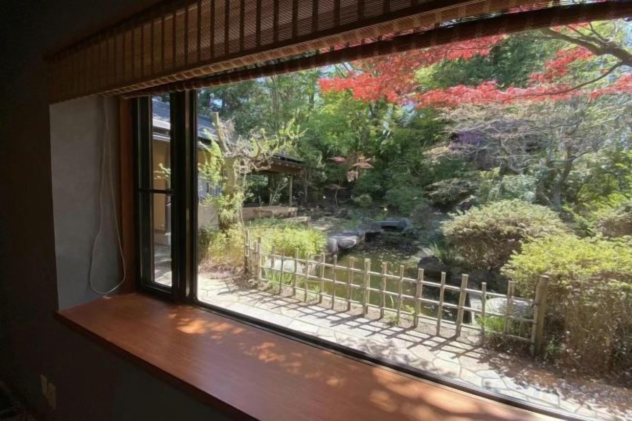 A view through a window with a wooden ledge, a bamboo blind, and a garden with a wooden fence and various plants.