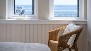 Een kamer met een bed, een rieten stoel en uitzicht op de oceaan door twee ramen.