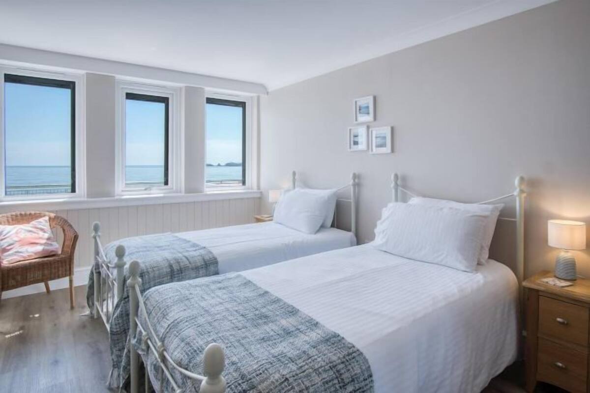 Une chambre avec deux lits, un fauteuil en osier, une table de chevet avec une lampe, et une vue sur la mer grâce à de grandes fenêtres.