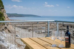 Une table en bois, avec deux verres à vin et une bouteille de vin, donnant sur une plage séparée par une clôture en fil de fer.