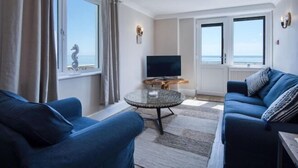 Een woonkamer met een bank, een rond glazen bijzettafeltje, een flatscreen televisie en een seahorse decoratie op de vensterbank.
