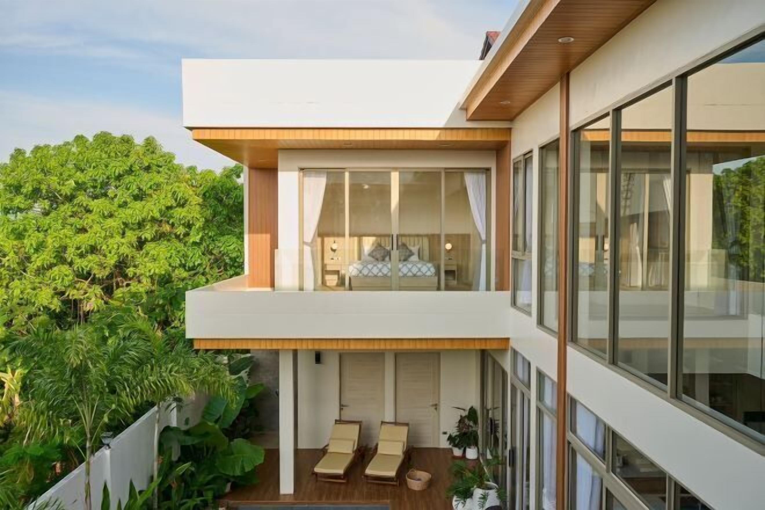 A modern two-story house with a balcony, large windows, and a view of lush greenery.
