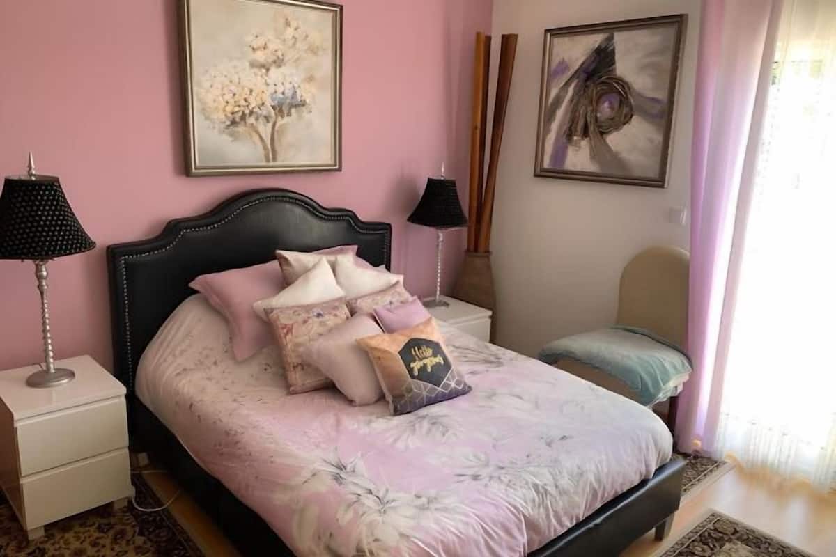 A bedroom with a bed, two bedside tables, a nightstand, a window with curtains, and two wall-mounted lamps.