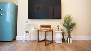 Een woonkamer met een flatscreen televisie aan de muur, een houten bijzettafeltje, een wit bijzettafeltje, een klein wit plankje en een blauwgroene koelkast, plus een potplant.