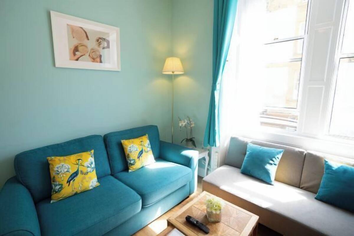 A teal-colored sofa with a remote control and a small plant on a wooden table, a lamp, and a framed picture on the wall.
