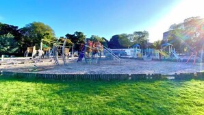Een speeltuin met diverse speeltoestellen, waaronder glijbanen en schommels, omringd door gras en bomen.