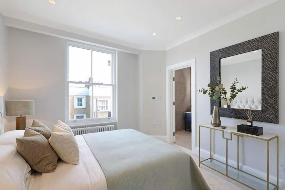 A bedroom with a large bed, a bedside table with a lamp, a window, and a mirror.