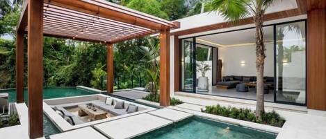 A modern outdoor pool area with a wooden pergola, lounge seating, and a glass-enclosed living space.