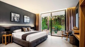 A modern bedroom with a large bed, a nightstand, a lamp, and a view of a lush garden through a sliding glass door. - Villa Oasis by BaliSuperHost (Bali)