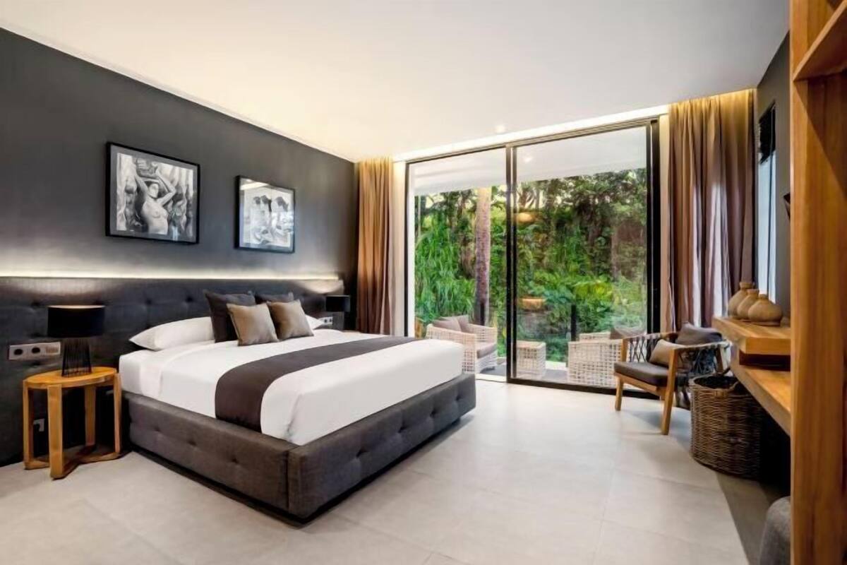 A modern bedroom with a large bed, a nightstand, a lamp, and a view of a lush garden through a sliding glass door.