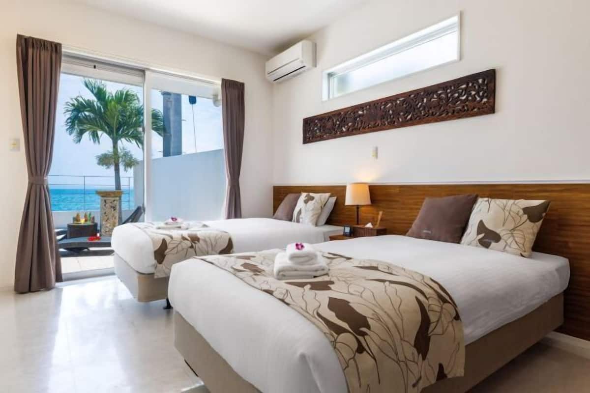 A hotel room with two beds, a large window with a view of palm trees and the sea, and a decorative wall piece above the beds.