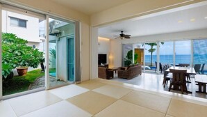 A spacious indoor area with a large sliding glass door leading to a balcony with a view of the ocean. There is a ceiling fan, a seating area with a table, and a TV mounted on the wall. - Coldio Marina Kin (Kin)