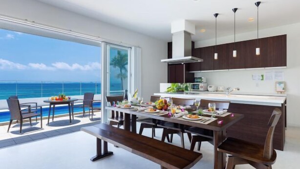 A modern kitchen with dark cabinetry and pendant lighting, a dining area with a view of the ocean, and a poolside seating area. - Coldio Marina Kin (Kin)