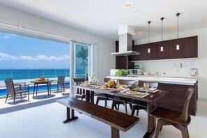 A modern kitchen with dark cabinetry and pendant lighting, a dining area with a view of the ocean, and a poolside seating area. - Coldio Marina Kin (Kin)