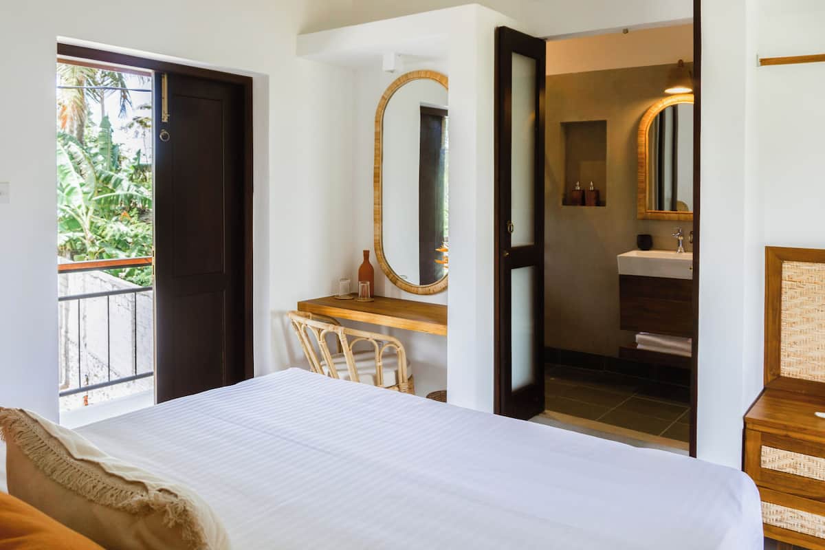 A hotel room with a bed, a desk, a chair, a bathroom with a mirror, and a view of greenery outside.