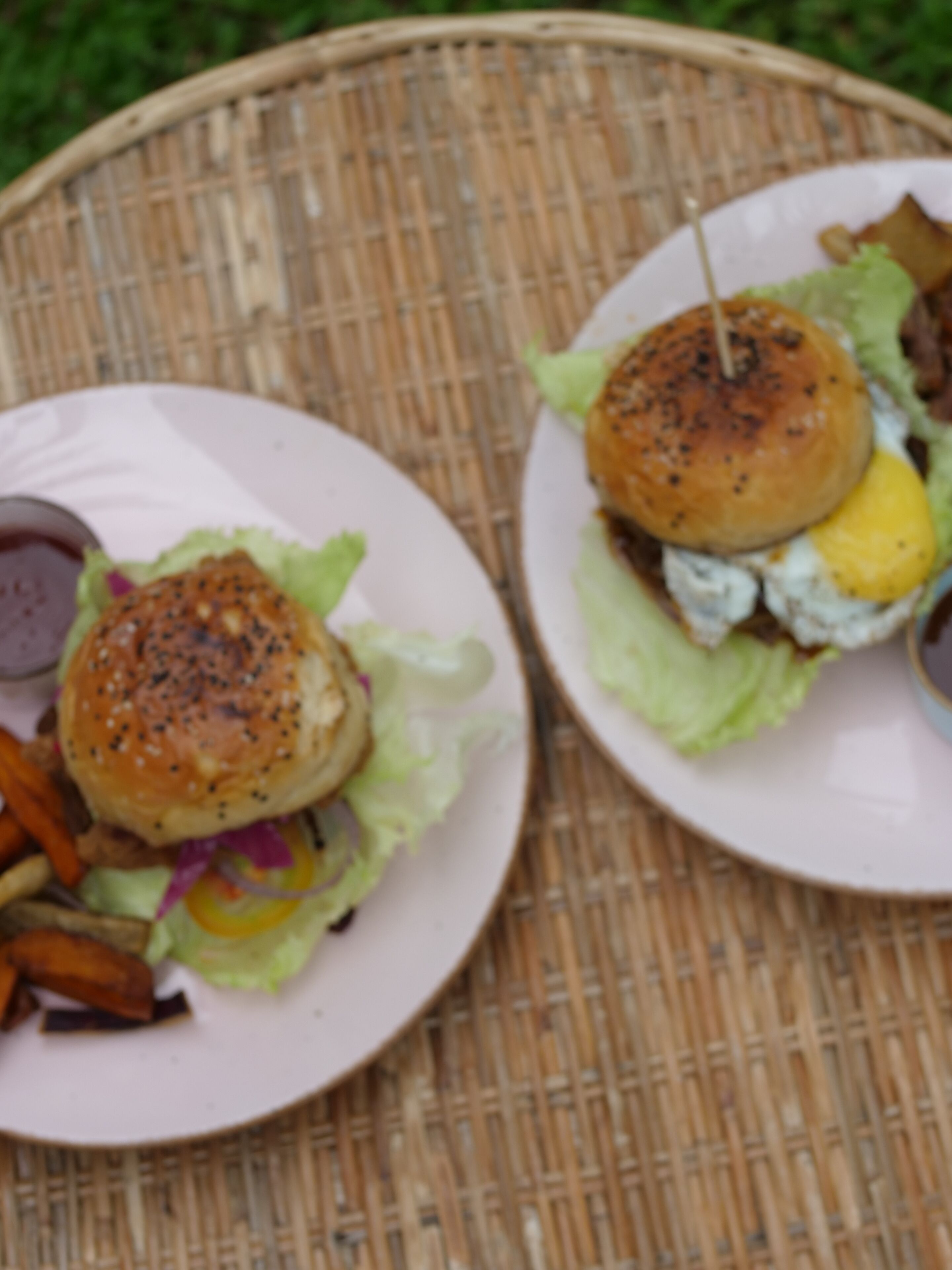 Two burgers on white plates with lettuce and a bun, placed on a woven basket.