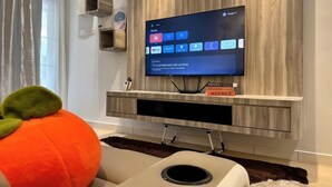 A modern living room with a flat-screen TV mounted on a wooden wall, a plush orange sofa, and a small shelf with books. - Austin Landed Honey Nest 10pax (Tebrau)