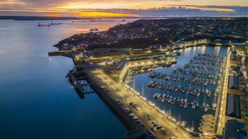 An aerial view of a coastal marina with boats docked, a pier, and a cityscape in the background.