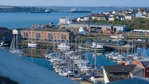 A marina with boats docked, a large building with a flat roof, and a residential area in the background. - The Penthouse - 1 Bedroom Penthouse - Milford Haven (Milford Haven)