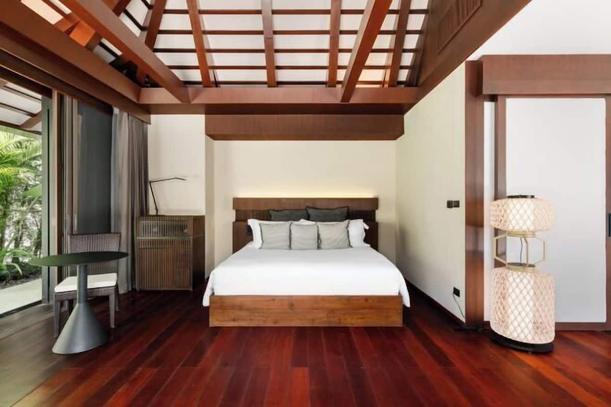 A modern bedroom with a wooden headboard, a bed with white bedding, a round table with a chair, and a floor lamp.
