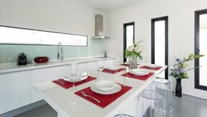 Une cuisine moderne avec des meubles blancs, un îlot central agrémenté de sets de table rouges et un coin repas avec des tabourets de bar transparents.