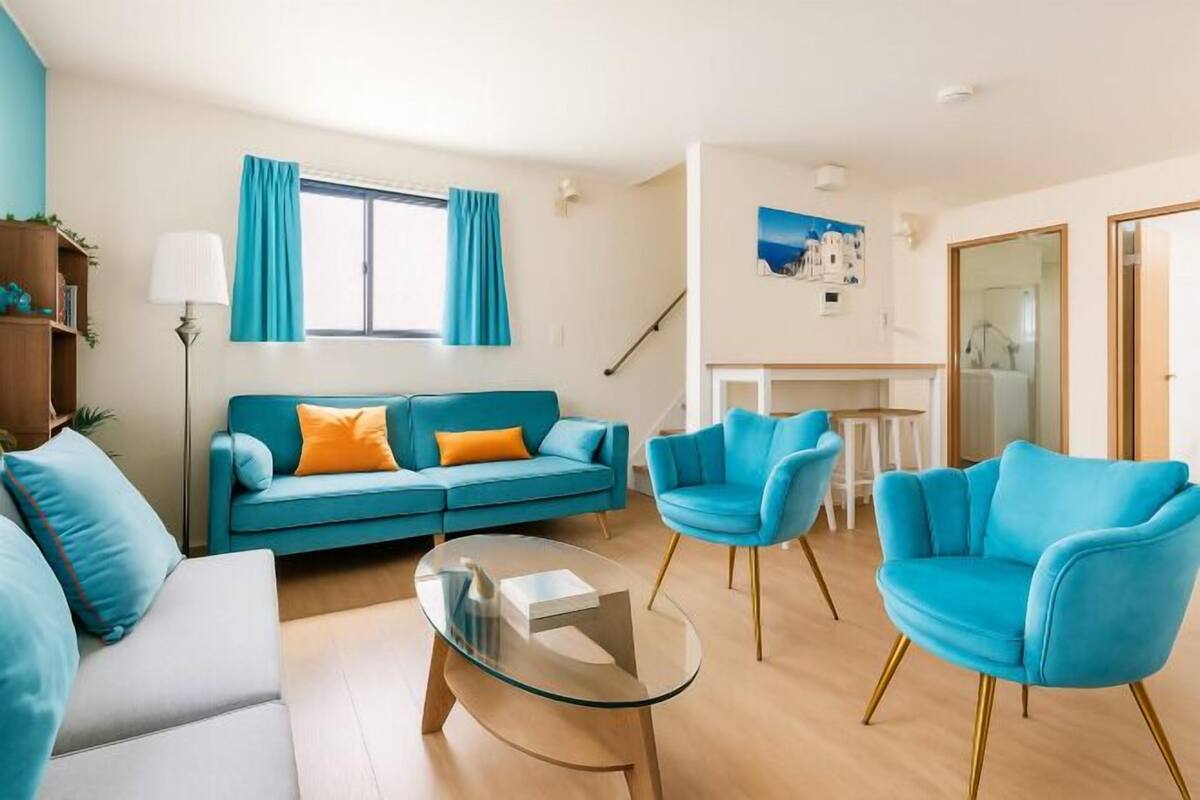 A modern living room with a teal sofa, matching armchairs, a glass coffee table, and a staircase leading to a dining area.