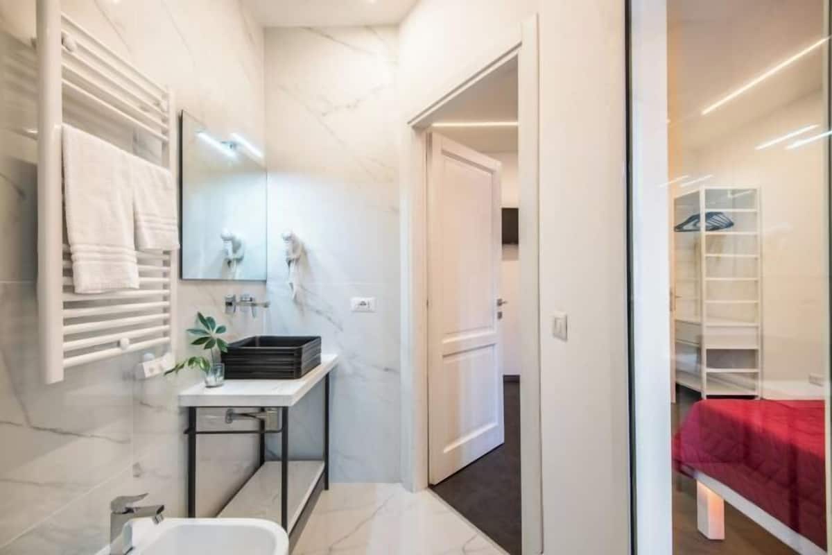 A modern bathroom with a white sink, mirror, and towel rack. A door leads to a bedroom with a red bedspread.