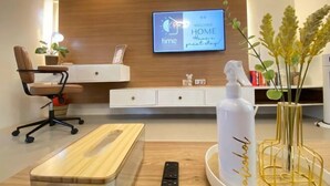 A modern hotel room with a wooden desk, a remote control, a bottle of alcohol, a clock displaying 'time HOME welcome', and a plant. - Time To Sleep — Casa Ala Una (Butuan)