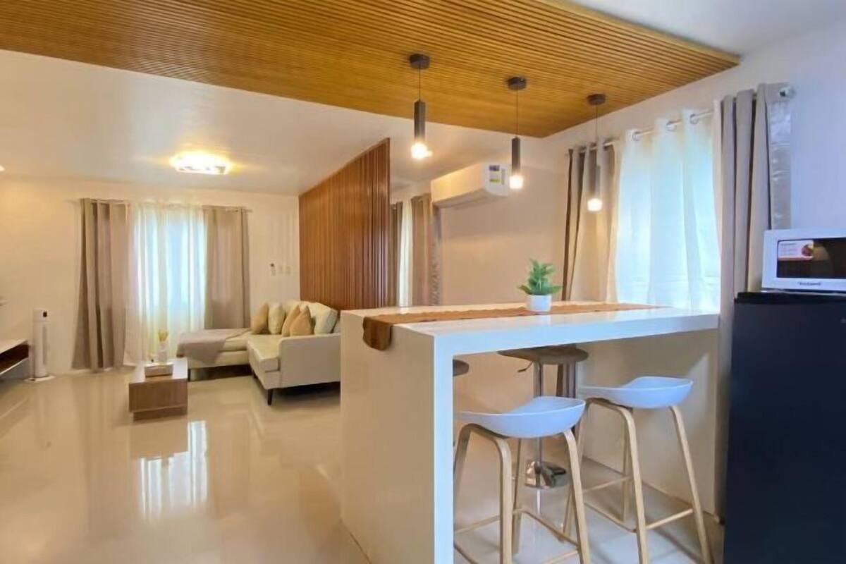 Una sala de estar moderna con un sofá, un comedor con taburetes, techo de madera y un televisor de pantalla plana.