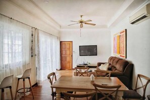 Un salon comprenant une table à manger en bois, des chaises, un canapé marron, un ventilateur de plafond et un téléviseur à écran plat fixé au mur.