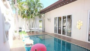 A swimming pool with a pink flamingo float, surrounded by palm trees and a patio area with white chairs.
