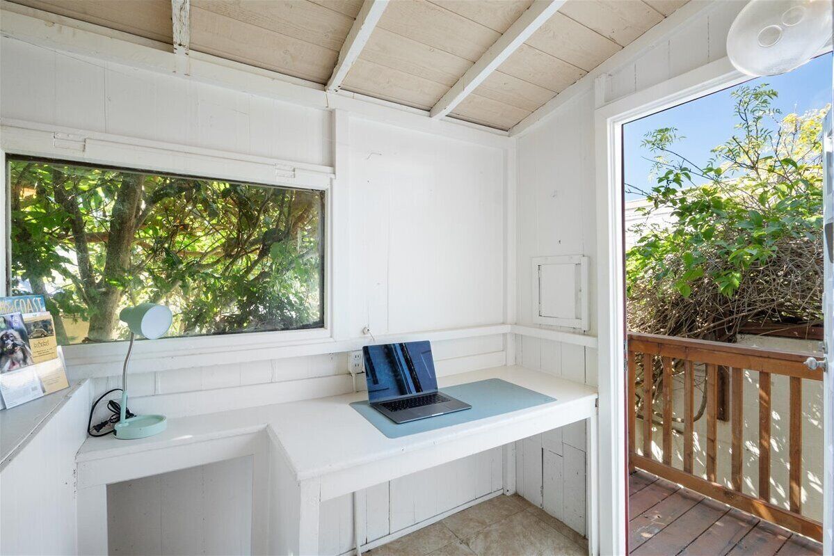 Un petit espace de travail clos, avec un ordinateur portable posé sur un bureau blanc, une fenêtre donnant sur des arbres et une terrasse en bois à l’extérieur.