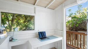 Un piccolo spazio di lavoro isolato con un laptop su una scrivania bianca, una finestra che si affaccia su alberi e un patio in legno all'esterno.