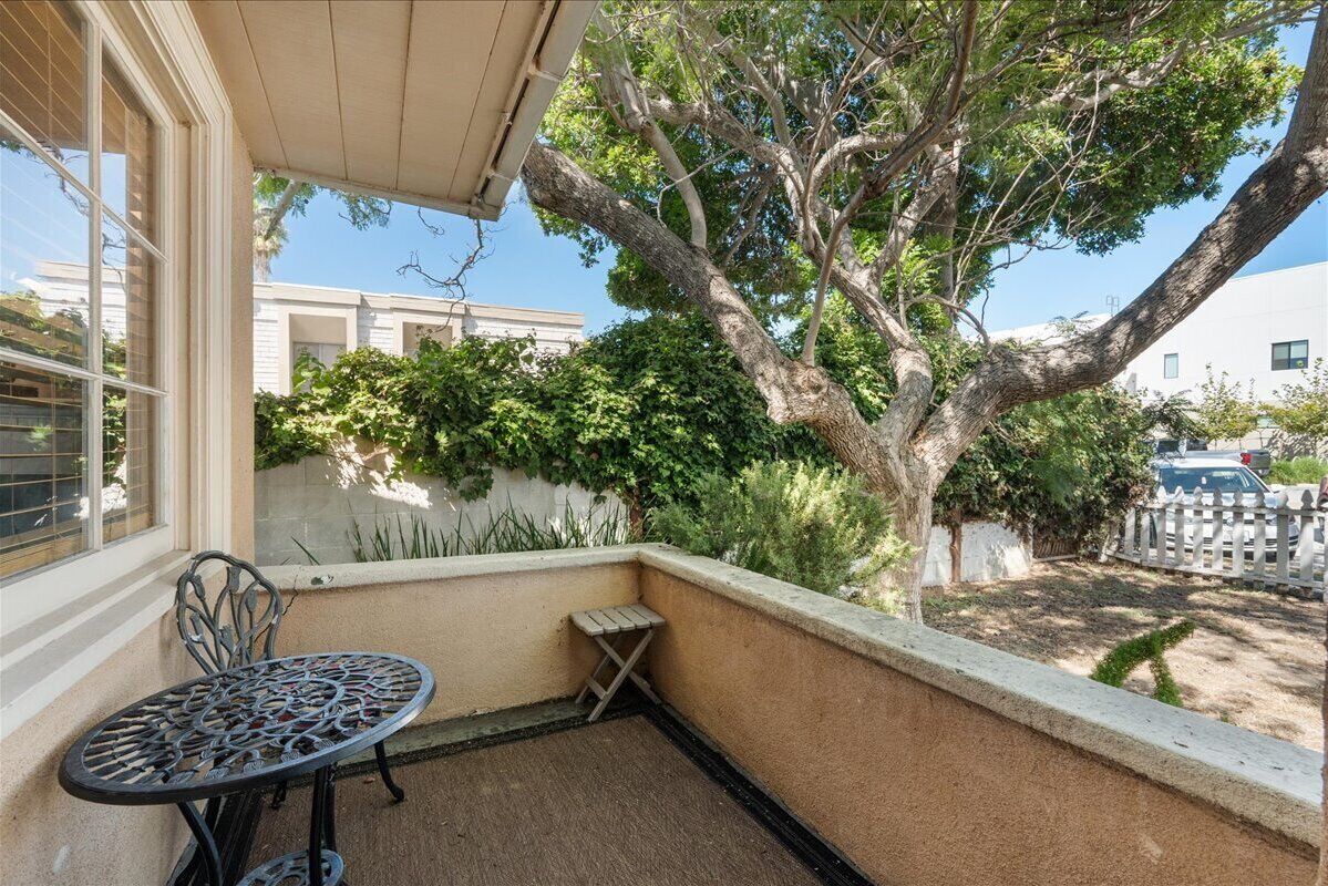 Une petite terrasse extérieure avec une table en métal et un tabouret en bois, entourée d’un petit muret et d’un grand arbre.
