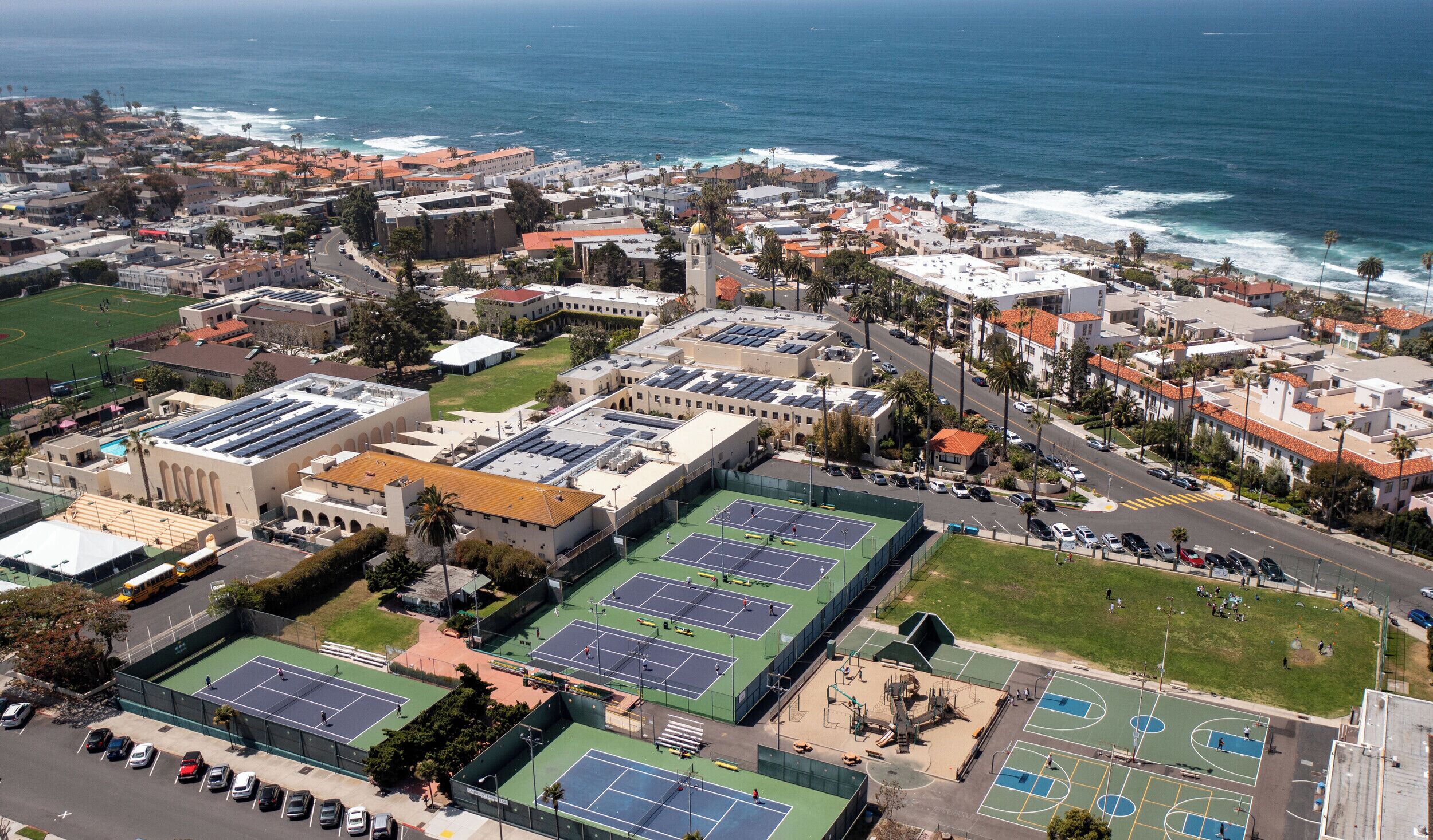 Une vue aérienne d’une ville côtière, avec un vaste complexe sportif comprenant des courts de tennis et de basketball, entouré de bâtiments résidentiels et d’une plage.
