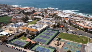 Vista aerea di una città costiera con un complesso sportivo in evidenza, dotato di campi da tennis e canestri da basket, circondato da edifici residenziali e una spiaggia.