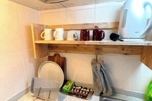Une cuisine avec une étagère en bois où sont posées des tasses et une bouilloire, une planche à découper et un porte-serviettes avec une serviette.