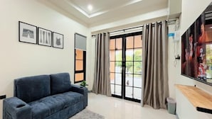 A modern living room with a dark blue sofa, a flat-screen TV mounted on the wall, and a large window with curtains. - DelZiaNa Bara Villa Pakbara Satun (La-ngu)