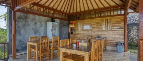 Un área de comedor exterior de madera con techo de paja, un refrigerador y una pared de piedra.