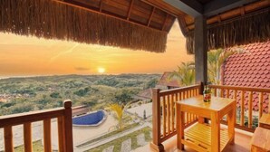 A wooden balcony with a table and chairs, overlooking a landscape with a pool and greenery. - Sebuluh Sunset Hill (Bali)