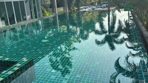 Una piscina con pavimento piastrellato, circondata da vegetazione e un edificio con ampie finestre.