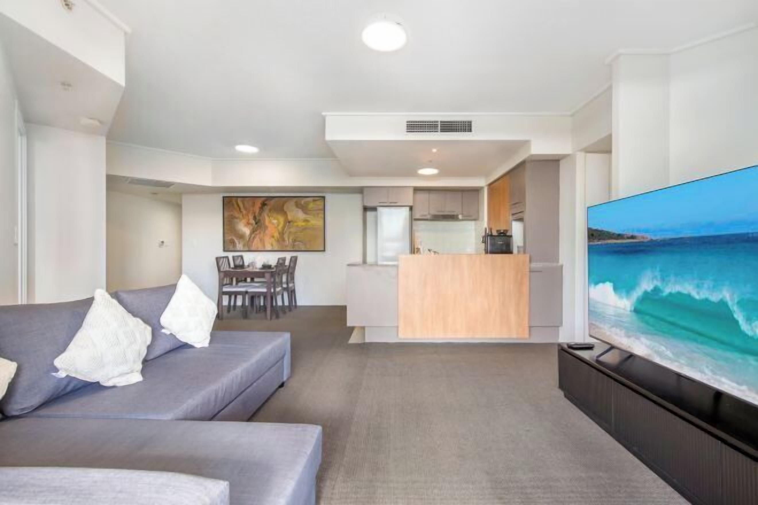 A modern living room with a grey sofa, a flat-screen TV, and a dining area in the background.