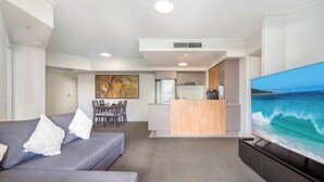 A modern living room with a grey sofa, a flat-screen TV, and a dining area in the background. - Arcade in the Sky - Views, Games & Vibes (Brisbane)