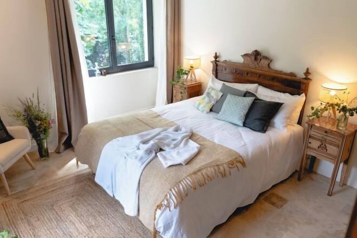 Un lit bien fait, avec une tête de lit en bois, deux tables de chevet avec des lampes, une chaise et une fenêtre avec des rideaux.
